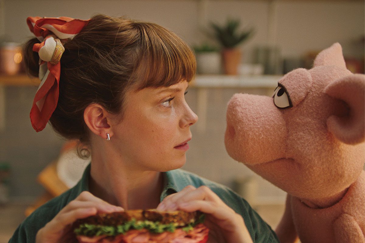 woman holding sandwich looking into a pig puppets eyes