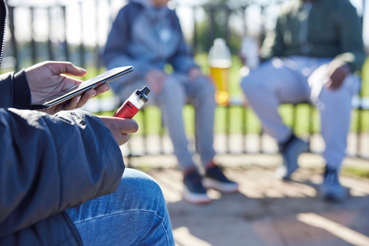 A youth holds their phone in one hand and a vaping device in their other hand with two young people blurred out in the background sitting on a bench with a plastic bottle between them.