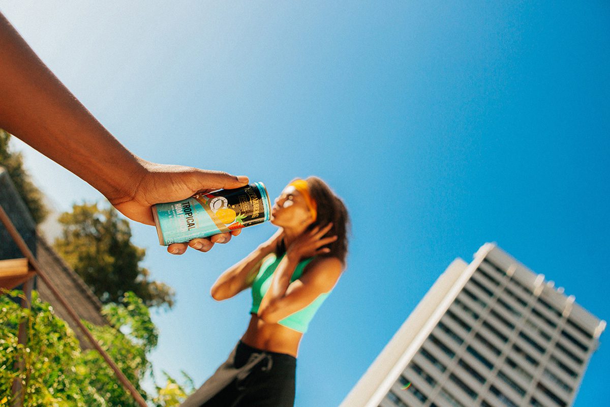 A hand holding a can of Kopparberg's new Tropical Rum Punch cocktail in the foreground makes it appear as though a woman running in the background is drinking from the can through a forced perspective.