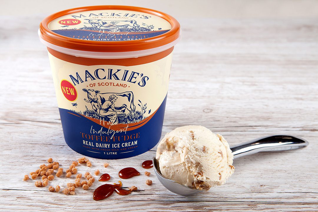 New Mackie's of Scotland Toffee Fudge ice cream tub with a scoop of the ice cream on a spoon next to it with pieces of fudge and toffee sauce.