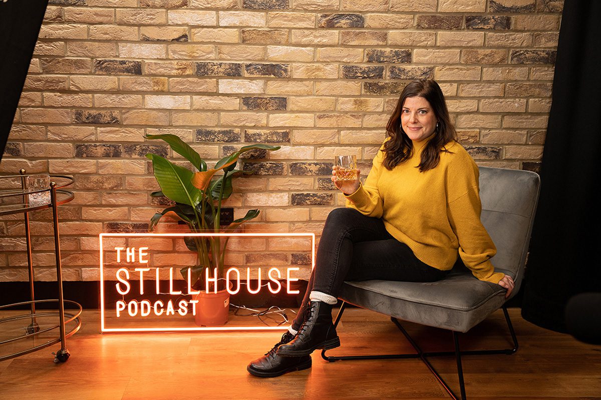 Becky Paskin of Edrington's The Stillhouse Podcast posing with a dram of whisky and a neon sign that says The Stillhouse Podcast.