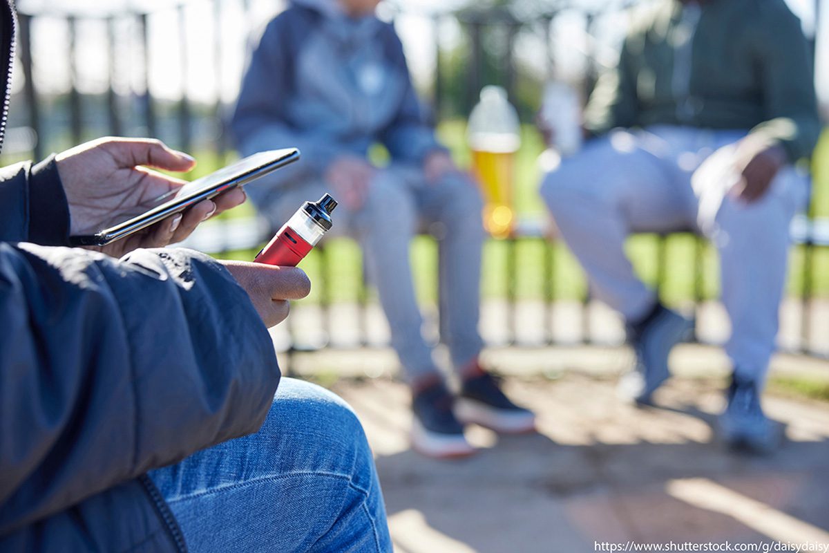 A young person sitting with a vape device in one hand and their phone in the other sits in the foreground with two others sitting blurred out in the background.Photo credit: https://www.shutterstock.com/g/daisydaisy