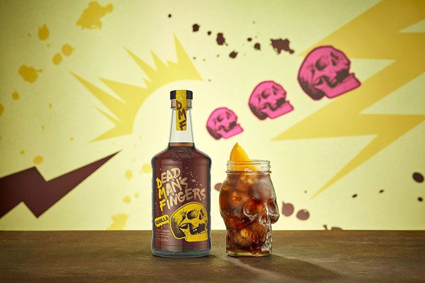 Dead Man's Finger's new Vanilla Rum bottle sits next to a skull shaped glass filled with a drink containing the rum set against a yellow wall that has three pink skulls on it.