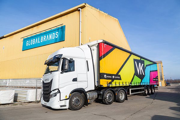 A HGV stands in front of a yellow warehouse with the Global Brands sign on it, the HGV includes a colourful truck with the VK logo across it.