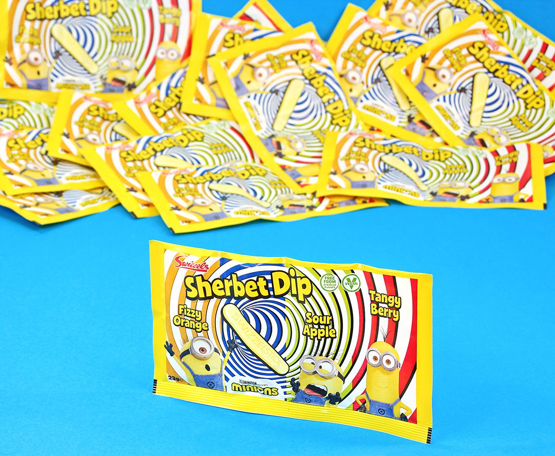 A single pack of Swizzels Minions Sherbet Dip stands in front of multiple packets of the sweet that have been scattered across a blue background.