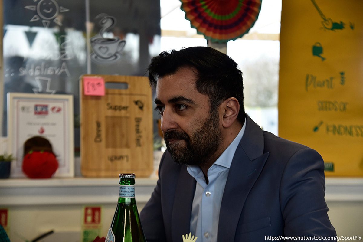 Scotland's first minister Humza Yousaf sits in a cafe with signs behind him with the top of a glass bottle visible in front of the first minister.