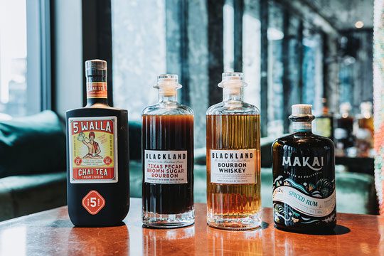 Bottles of 5 Walla Chai Liqueur, Blackland Texas Pecan Brown Sugar Bourbon, Blackland Bourbon Whiskey and Makai Spiced Rum sit in a row on top of a wooden table with a blurry green couch in the background.