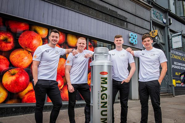 Scottish curling team Team Mouat stand next to a novelty size bottle of Magnum Scotch whisky cream liqueur outside of a Co-op store.