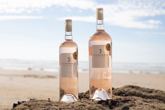 Two bottles of Sea Change Provence Rose sit on top of a rock with a beach in the background of the photo.
