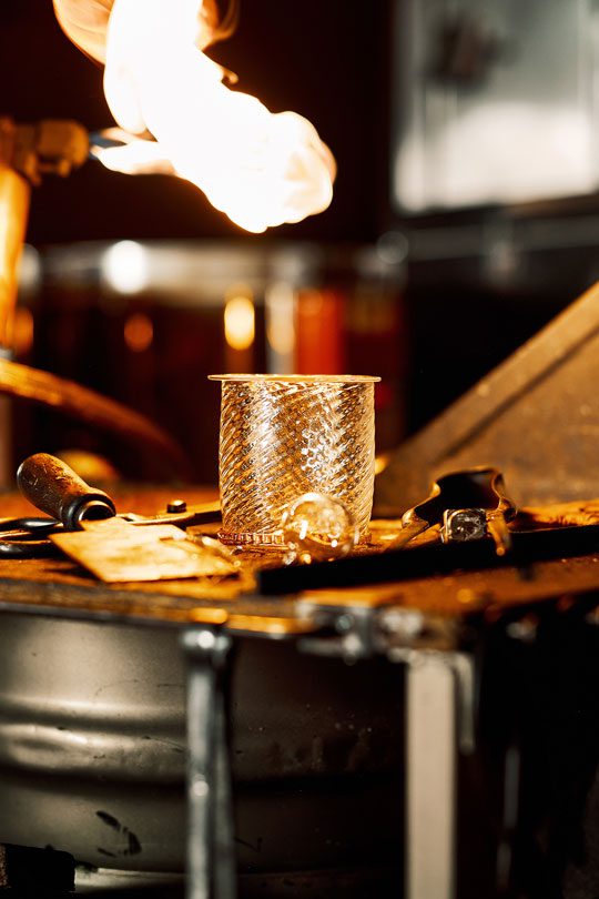 A prototype whisky glass sits on a workbench with tools on either side of it and a flame above it.
