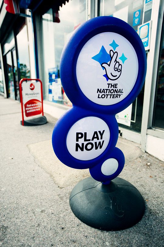 A sign for the National Lottery stands outside of the front of a shop with a Moneygram sign in the background.