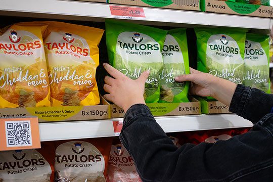 Taylors Potato Crisps flavour variants sit on a shelf as a customer reaches out to pick up a green packet of Pickled Onion flavoured crisps