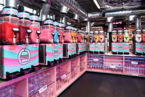 A row of Tubbees branded slush machines with different flavours all sit on top of neon pink shelving units with plastic cups underneath.