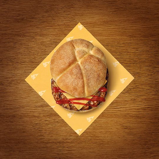 A Rollover cheeseburger with tomato ketchup sits and yellow napkin which has cheese symbols on it sit on top of a wooden table.