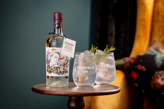 A bottle of the new collaborative gin from Edinburgh Gin and Suzy Eddie Izzard sits next to two Edinburgh Gin marked glasses filled with a gin and tonic. All of this sits on a wooden circular table with a blue wall in the background.
