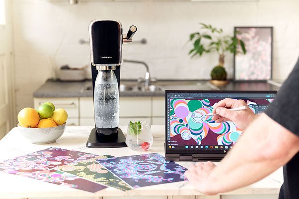 A person is designing new artwork on a tablet screen with a Sodastream Art machine on the table next to the person.