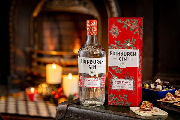 A bottle of Edinburgh Gin Christmas sits next to a packaging box of Edinburgh Gin Christmas gin in front of a festive scene with a fireplace with candles on it in the background. The gin sits on a table with a cheese and crackers spread.