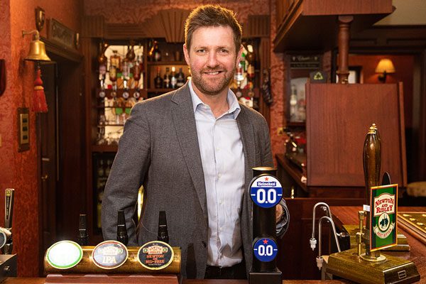 A man stands behind a bar highlighting the Heineken 0.0% beer tap. The bar itself is wooden with three other beer taps across it with a bell in the background to the left.