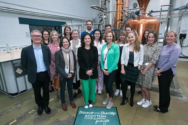 Group of people at Lind & Lime Distillery to launch the start of Scottish Food & Drink Fortnight with a doormat at the front that says: "Discover what's on your doorstep. Scottish Food & Drink Fortnight."