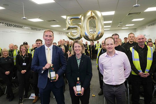 (Left to right) Ewan Andrew, president, global supply chain & procurement and chief sustainability officer at Diageo, Jenny Gilruth MSP, cabinet secretary for education and skills, and Stuart Galbraith, Diageo Leven site director, stand in front of crowded room of employees at Diageo Leven. Two golden balloons are behind the three, one a 5 and the other a 0 to mark 50 years of the Leven site. 