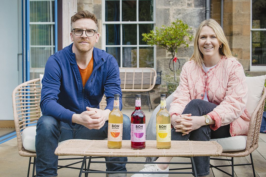 Nathan Burrough, director of Bon Accord Soft Drinks, and Karen Knowles, managing director of Bon Accord Soft Drinks, sit on a couch next to each other with three bottles of Bon Accord soft drink variants on the table in front of them.