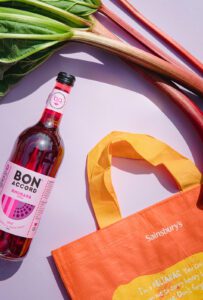 A bottle of Bon Accord Rhubarb Soda lies on its side next to a Sainsbury's branded bag and sticks of rhubarb above it against a violet coloured background.
