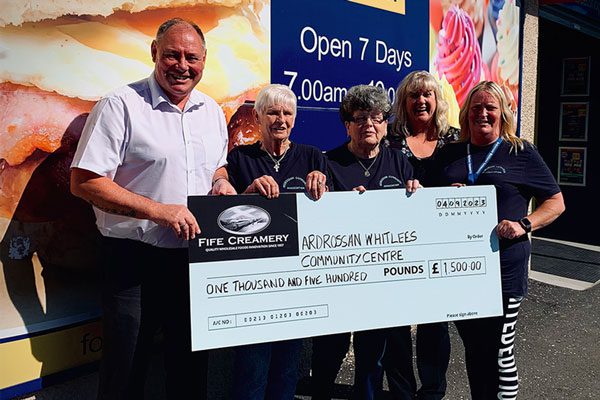 Five people stand in front of a store holding a cheque, the man on the far left is wearing a white shirt with four women all dressed in dark colours to the right of him. They hold a cheque with the Fife Creamery logo on the top right hand corner with the cheque stating: ARDROSSAN WHITELEES COMMUNITY CENTRE, ONE THOUSAND AND FIVE HUNDRED POUNDS