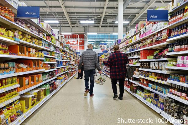 Supermarket aisle with lines of ambient foods stocking. People walk down the middle of the aisle holding shopping baskets and having a conversation.