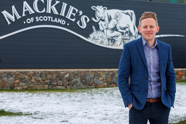 Stuart Common, managing director for Mackie's of Scotland, stands in front of a grey wall with the Mackie's of Scotland logo behind him which depicts a woman milking a cow.