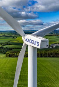 A Mackie's of Scotland wind turbine with the Mackie's logo on it. The turbine stands above fields of green with a blue sky and clouds.