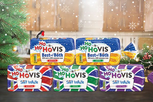 Image depicting the new Ho Ho Hovis seasonal packs for Christmas. The five limited edition packs sit stacked together on a table in a kitchen, with Christmas tree branches to the left of the packs and a holly wreath to the right. Snow is falling from above the packs.