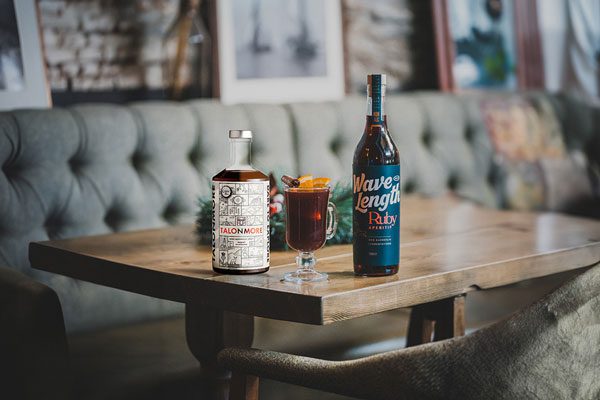 A bottle of Talonmore and a bottle of Wave Length Ruby Aperitif both sit on a wooden table with a TalonWave cocktail in between the two of them. There is a grey couch in the background with framed pictures above the couch.