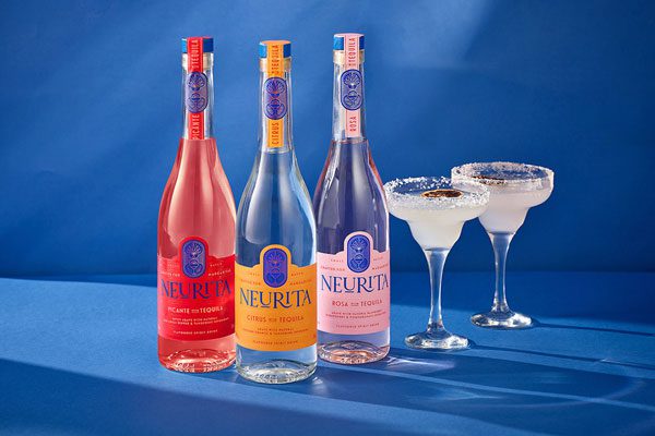 Three bottles of Neurita Tequila stand next to each other against a blue blackground. (From left to right) Neurita Picante, Neurita Citrus, Neurita Rosa. To the right of Neurita Rosa are two margarita glasses filled with the cocktail with a salt rim on the glass.