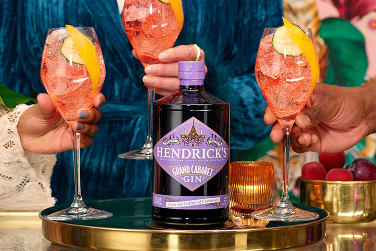 A bottle of Hendrick's Grand Cabaret gin sits on a table as three people hold cocktails containing the gin around it.