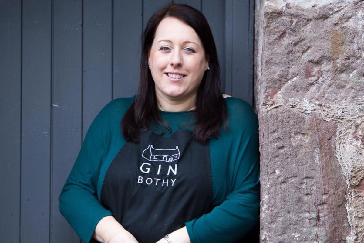 Kim Cameron, owner of Gin Bothy, stands in front of a wooden door in a dark green top wearing a Gin Bothy branded black apron.