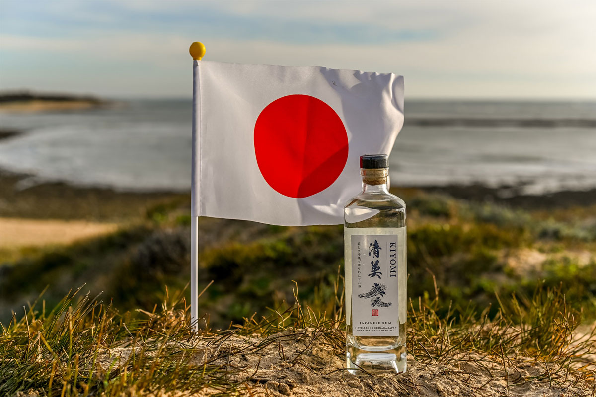 A bottle of Kiyomi Rum stands in front of a Japanese flag blowing in the wind which has been planted into the grassy ground all of which overlooks a beach from atop a cliff.