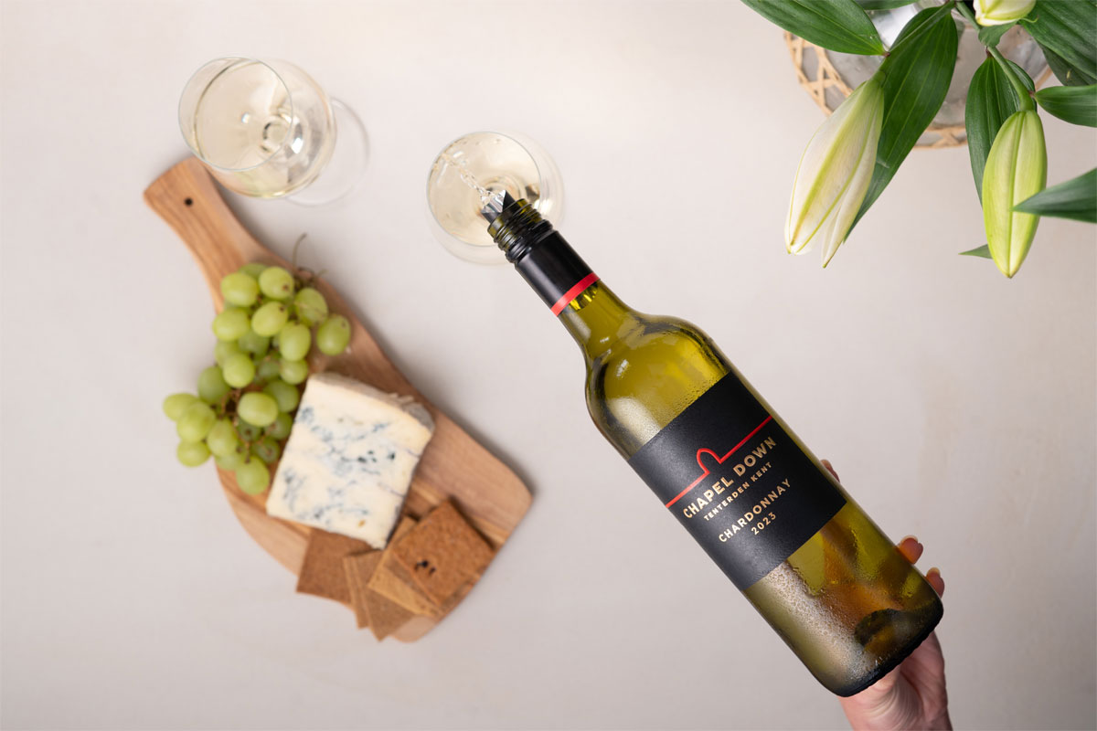 A hand pours out Chardonnay wine from a bottle of Chapel Down Chardonnay 2023 into a glass against a white background with a already filled wine glass and cheeseboard with white grapes next to the glasses.