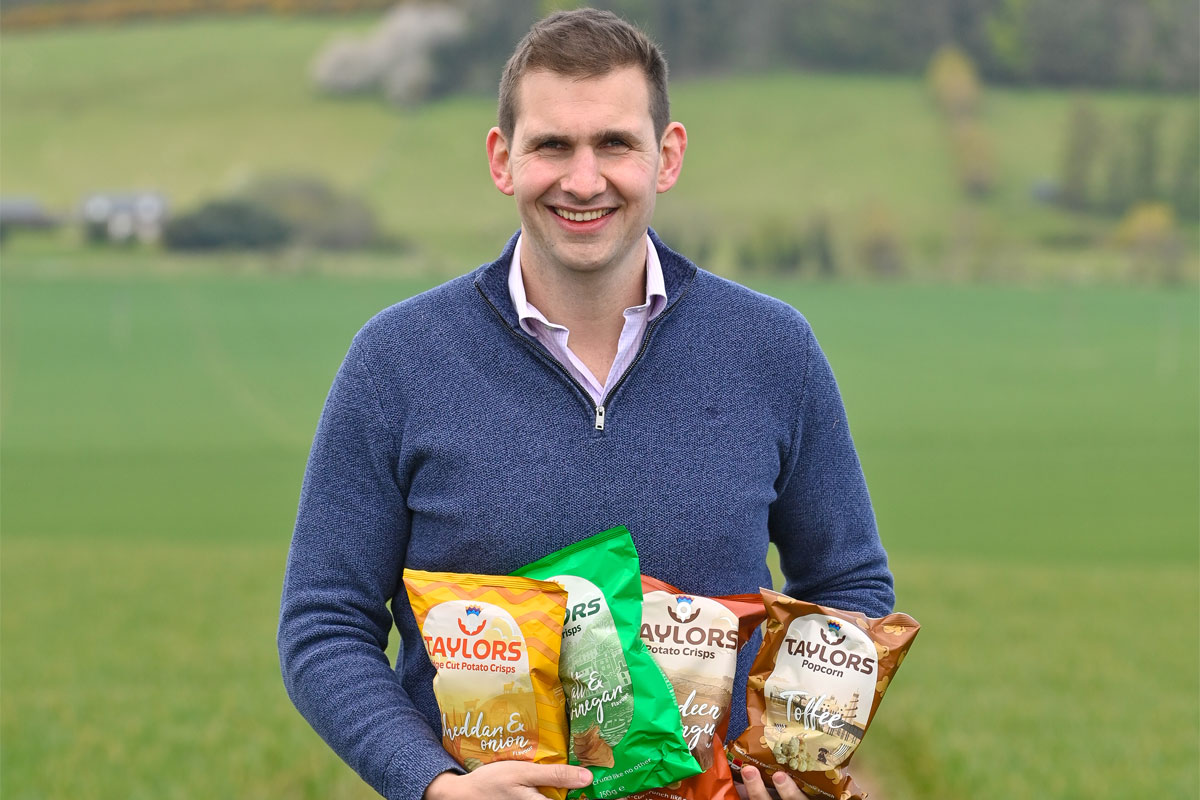 James Taylor, MD at Taylors Snacks, stands holding packs of Taylors Snacks crisps against a green field.