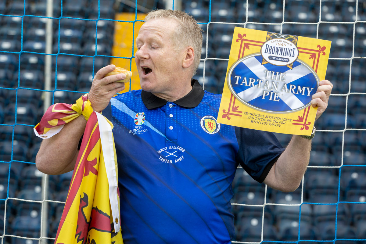A man is seen going to eat a pastry pie wearing the Scotland Football Team's top and holding a Lion Rampant flag as well as a card with the branding for Browning's The Tartan Army Pie.