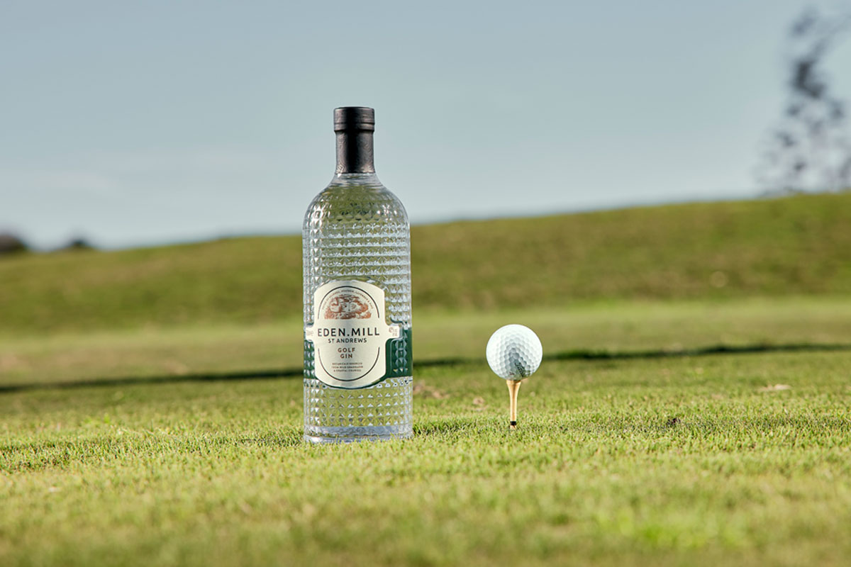 A bottle of Eden Mill Golf Gin sits next to a golf ball on a tee in the middle of a green.
