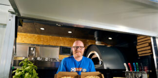 Gary McLean, Scotland's national chef, stands in a kitchen with a pizza in each hand with the pepperoni on the pizzas spelling out 90,000.
