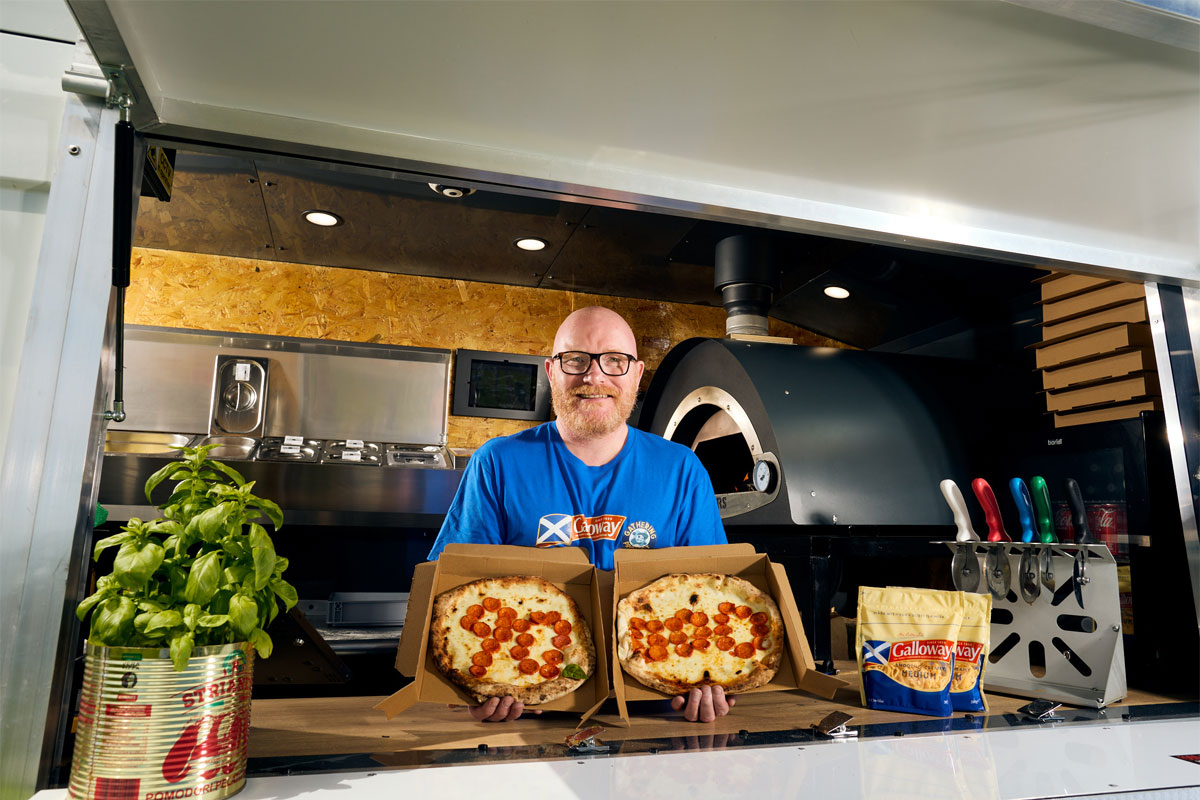 Gary Maclean, Scotland's national chef, stands in a kitchen with a pizza in each hand with the pepperoni on the pizzas spelling out 90,000.