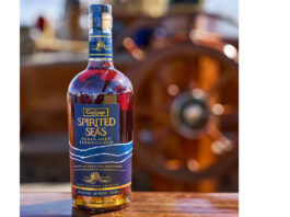 New Goslings Rum docks in the UK A bottle of Goslings Spirited Sea sits on a wooden bannister of a ship with a ship's wheel blurred in the background.