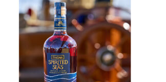A bottle of Goslings Spirited Sea sits on a wooden bannister of a ship with a ship's wheel blurred in the background.