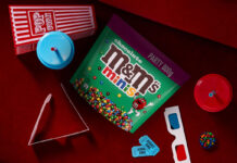 A packet of M&Ms Minis Party Size sits on a cinema seat with a popcorn box, juice cups, cinema tickets and 3D glasses around it.