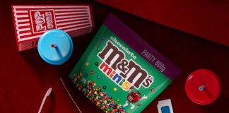 A packet of M&Ms Minis Party Size sits on a cinema seat with a popcorn box, juice cups, cinema tickets and 3D glasses around it.