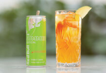 A can of Red Bull Curuba Summer Edition is next to a glass filled with the energy drink on a table with greenery blurred in the background.