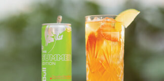 A can of Red Bull Curuba Summer Edition is next to a glass filled with the energy drink on a table with greenery blurred in the background.