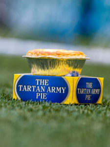 A Brownings The Tartan Army Pie sits on top of its box packaging on a football field.