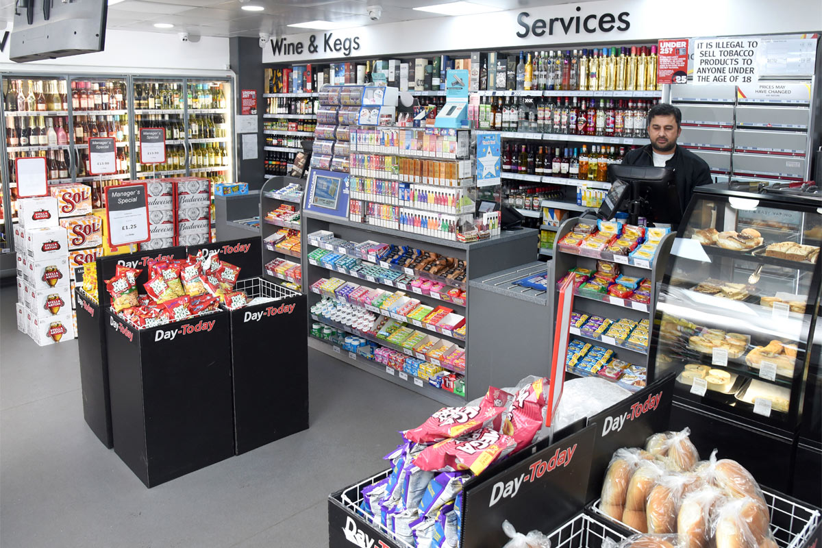 A Day-Today store floor with the counter area, product bins close to the checkout and a staff member behind the till showing products affected by inflation.